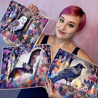 Woman with pink hair holding three large art prints of nocturnal animals surrounded by vibrant foliage.