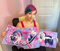 A woman with pink hair displays three colorful paintings of a raccoon, skunk, and opossum from the Trash Animals - Fine Art Print Bundle in a home setting.